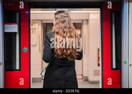 Una donna sta in piedi in una stazione della metropolitana guardando verso le porte aperte del treno mentre aspetta l'arrivo del treno successivo, mostrando la routine quotidiana di Foto Stock