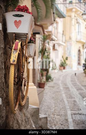 Bicicletta gialla decorativa con cesti di fiori su una parete lungo un vicolo acciottolato in una città italiana meridionale, che riflette il fascino mediterraneo e lo stile di vita rilassato. Foto Stock