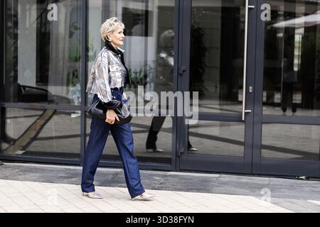 Donna matura e sicura che cammina all'aperto nella moderna città indossando giacca argento metallizzato, pantaloni in denim e tacchi. Elegante modello di donna anziana con Foto Stock