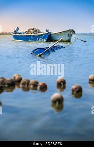 Vista ravvicinata dei galleggianti in sughero e di una rete sommersa, con un peschereccio bianco e blu ancorato a Etang de Canet, Francia Foto Stock