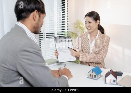 Un agente immobiliare discute i contratti immobiliari con un cliente, utilizzando modelli di abitazione e una calcolatrice per spiegare i termini. La scena evidenzia la negoziazione Foto Stock