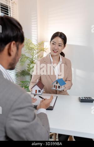 Un agente immobiliare discute i contratti immobiliari con un cliente, utilizzando modelli di abitazione e una calcolatrice per spiegare i termini. La scena evidenzia la negoziazione Foto Stock