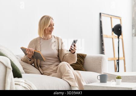 Una donna bionda si rilassa nel suo accogliente appartamento, godendosi momenti tranquilli con un libro e un telefono. Foto Stock