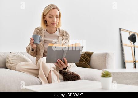Una donna bionda si rilassa a casa con abiti comodi mentre si gusta un libro e una bevanda calda. Foto Stock