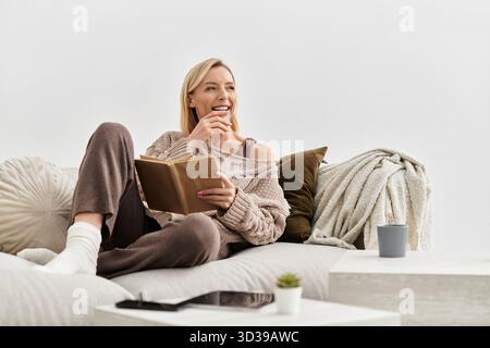Una donna bionda si rilassa sul suo divano, leggendo e mangiando spuntini nel suo accogliente appartamento. Foto Stock
