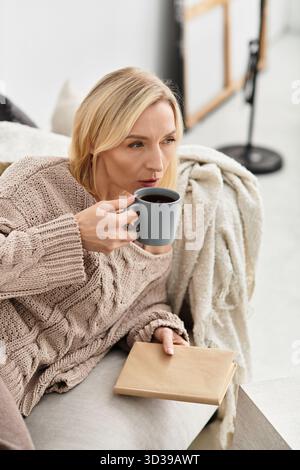 Una donna bionda si rilassa nel suo moderno appartamento, sorseggiando un caffè mentre è avvolta nel comfort. Foto Stock