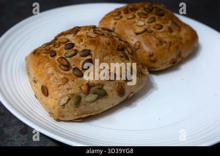 Due rotoli dorati ricoperti di semi di zucca sono disposti ordinatamente su un piatto bianco. Questi panini freschi sono perfetti per la colazione o come salutari Foto Stock