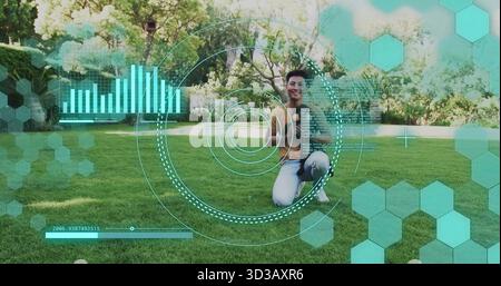 Inginocchiato uomo asiatico con la camicia di senape, pantaloni grigi che danno i pollici sul prato del parco, con HUD digitale Foto Stock