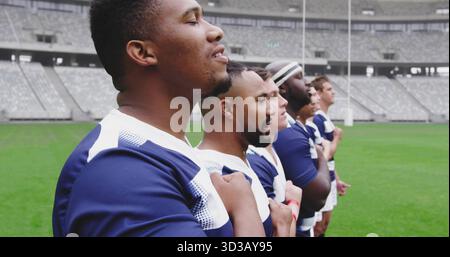 In piedi otto giocatori di rugby che tengono le mani giuste sopra il petto in campo con maglie navy, copia spazio Foto Stock