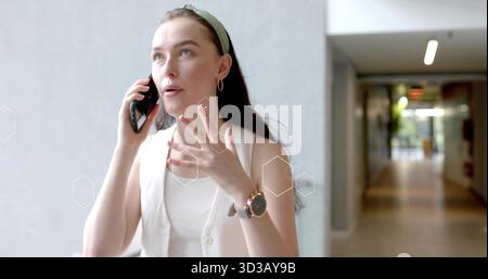 Donna che parla su smartphone in corridoio d'ufficio, indossa un giubbotto con grafica esagonale Foto Stock