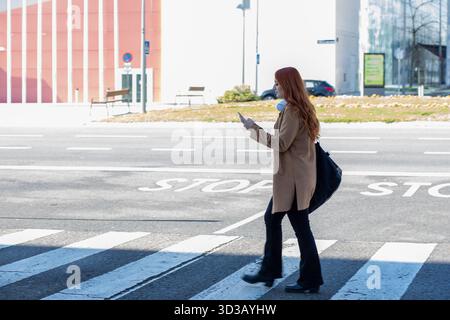 Una donna con lunghi capelli rossi cammina attraverso un incrocio in una città moderna, concentrata sul suo smartphone. Il tempo è limpido e soleggiato, evidenziando l'urba Foto Stock