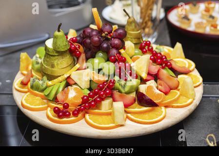 Cibo, frutta fresca assortita, tra cui uva, arance, pere e kiwi, splendidamente disposti su una tavola rotonda in legno per il catering Foto Stock