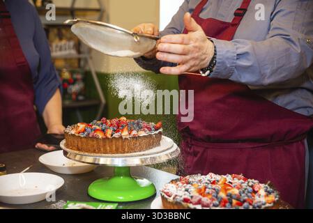 Il cibo, la persona in grembiule cosparge di zucchero a velo su una crostata di frutti di bosco appena preparata, aggiungendo un tocco decorativo finale in cucina Foto Stock