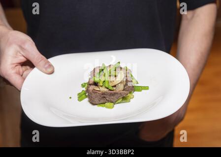Cibo, filetto di manzo cotto servito su un piatto bianco, condito con funghi saltati e asparagi tritati per una presentazione gourmet Foto Stock
