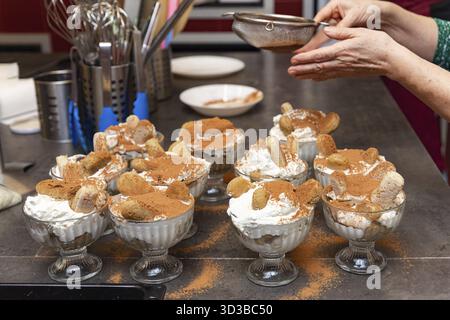 Cibo, più tazze in vetro di tiramisù fatti in casa sono ricoperte di cacao in polvere utilizzando un setaccio in un ambiente accogliente in cucina Foto Stock