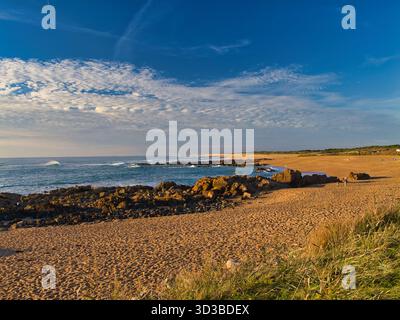 La calda luce del tramonto bagna un'ampia spiaggia sabbiosa e affioramenti rocciosi a Les Sables-Dolonne sulla costa di Vendea, Francia, con onde dolci e nuvole a motivi geometrici. Foto Stock