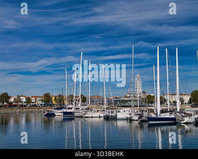 La Rochelle, Francia - 02 ottobre 2025: Barche a vela e yacht ormeggiati a la Rochelle, in Francia, con una ruota panoramica e un lungomare che si riflettono in acque calme sotto un cielo azzurro. Luce pomeridiana brillante e nitida. Foto Stock