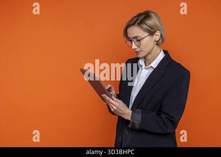 Donna seria che indossa una tuta nera in stile ufficiale che lavora online su un tablet isolato su sfondo arancione Foto Stock