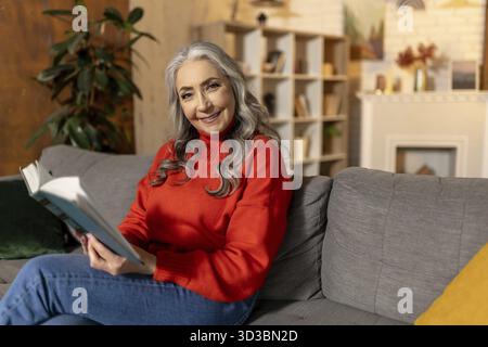 A casa. Bella donna anziana dai capelli lunghi seduta sul divano a casa Foto Stock