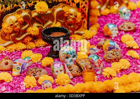 Vista ravvicinata dei teschi di zucchero messicani e del pane Pan de Muerto su un altare coperto da vivaci petali di calendula rosa e arancia. Foto Stock