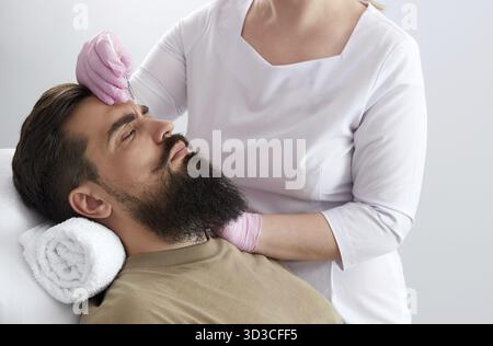 Primo piano di un uomo barbuto che riceve un'iniezione di bellezza alla clinica estetica. Medico iniettando il riempitivo anti-invecchiamento in bel viso maschile. Cosmetologia maschile, aesth Foto Stock