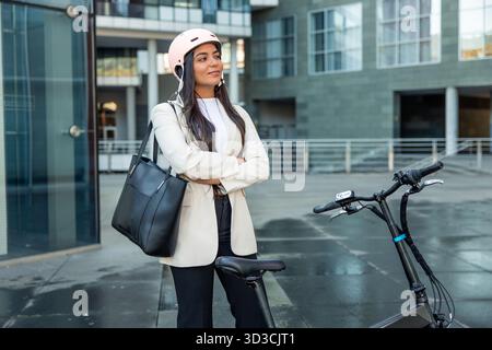 Donna d'affari latina in piedi in modo confidenziale accanto a una bici elettrica pieghevole, che indossa un casco e porta una borsa tote, che abbraccia spostamenti urbani sostenibili Foto Stock