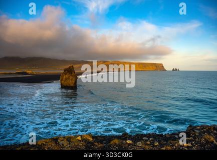 La spiaggia di Kirkjufjara presenta sabbia nera e formazioni rocciose uniche sotto un vibrante tramonto. La costa presenta onde dolci e montagne panoramiche Foto Stock