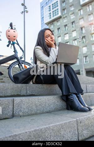 Donna d'affari latina seduta su scale di cemento con un portatile, che si riflette e guarda lontano mentre la sua bici elettrica pieghevole le aspetta accanto in un moderno fi Foto Stock