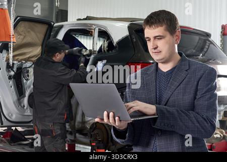 Manager che tiene in mano un notebook mentre si trova vicino a un'auto in un'officina di riparazione con meccanico che ripara l'auto in background Foto Stock