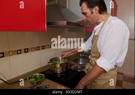 Un uomo con camicia bianca e grembiule mescola una pentola sul piano cottura, preparando la zuppa in una cucina contemporanea. Una ciotola laterale di green si trova nelle vicinanze, suggerendo fam Foto Stock
