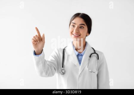 La giovane dottoressa in cappotto bianco punta allo spazio vuoto con un sorriso sul viso Foto Stock