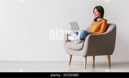 Gioiosa donna millenaria che studia online mentre è seduta su una poltrona con un laptop e cuffie Foto Stock