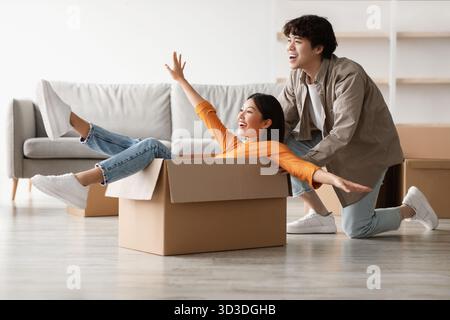 Una coppia asiatica giocosa si diverte a traslocare divertendosi in una scatola di cartone nella loro nuova casa Foto Stock