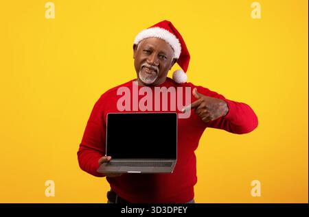 L'uomo nero senior che indossa un cappello di Babbo Natale punta felicemente su un notebook con schermo vuoto Foto Stock