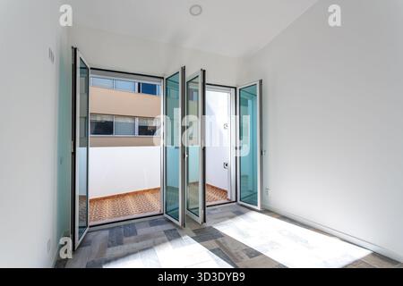 Spazio aperto al sole con balcone e elementi di design minimalista, sala luminosa con porte in vetro pieghevoli che conducono al balcone con legno Foto Stock