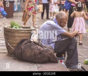 Chongqing, Cina - agosto 2019: Vecchio cinese stanco che riposa accanto alla sua grande borsa di tela sul marciapiede Foto Stock