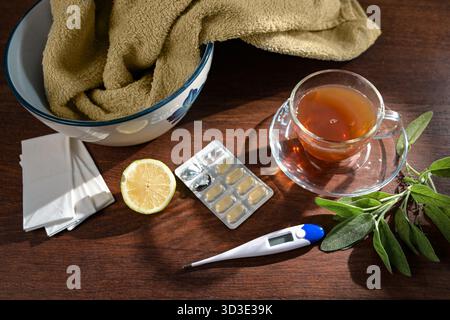 Cure domiciliari contro il raffreddore e l'influenza, tisane, salvia, limone, pillole, salviette e un recipiente con asciugamano per inalazione di vapore su un tavolo di legno, hea Foto Stock