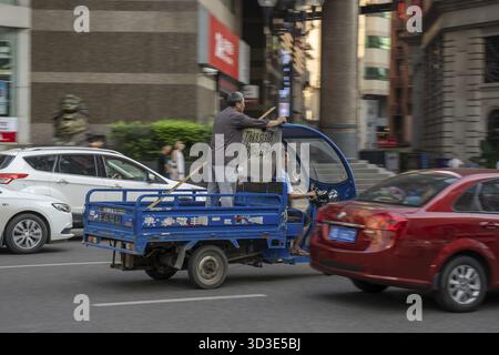 Chongqing, Cina - Agosto 2019 : l'uomo in piedi sul retro di un piccolo pick up risciò guida pericolosissima veloce sulla strada congestionata di Chongqing Foto Stock