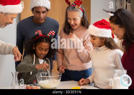 Famiglia variegata in cappelli per le vacanze che si riuniscono al banco della cucina mescolando la pastella in una ciotola di vetro Foto Stock