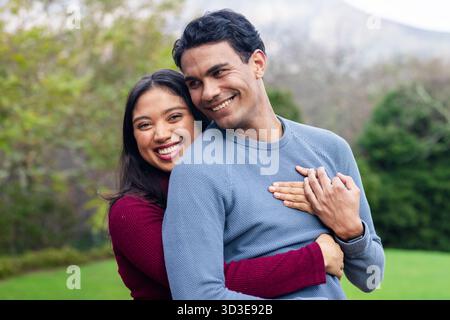 Coppia romantica che indossa maglioni che sorride e abbracciano nel verde del parco vicino agli arbusti e alle colline lontane Foto Stock