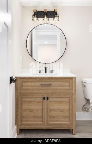 Un bagno moderno in stile fattoria con un ripiano bianco su un armadietto in quercia bianca, lampade nere e dorate sopra uno specchio circolare, e ta Foto Stock