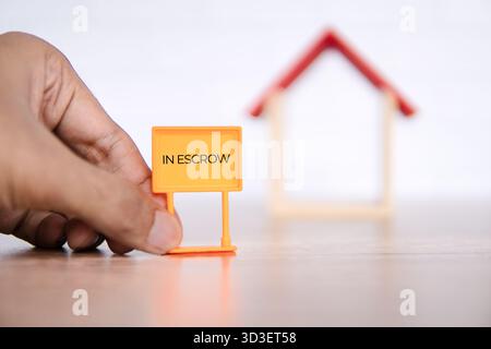 Una mano regge un cartello immobiliare "IN DEPOSITO", con una casa in miniatura sfocata sullo sfondo. Il processo di acquisto e vendita di immobili Foto Stock