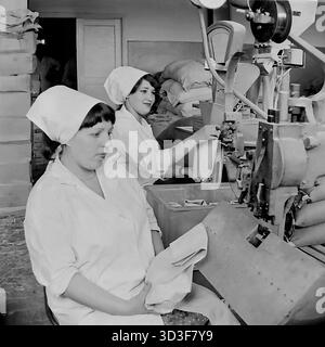 Un'altra vista del deposito in un negozio a Cherevkovka, Sloviansk, RSS Ucraina, durante gli anni '1970 Due lavoratrici in camice e velo bianchi azionano la macchina. In primo piano, una donna alimenta un sacchetto di carta in una macchina per sigillare o cucire. La seconda donna dietro di lei gestisce la stazione di rifornimento, con una tramoggia e bilancia sopra. Stanno elaborando merci sfuse per il negozio. Questa foto d'archivio mostra l'intenso lavoro "dietro le quinte" in un negozio sovietico. Questo è un momento di lavoro quotidiano nel pacifico Donbas prima della guerra. Foto Stock