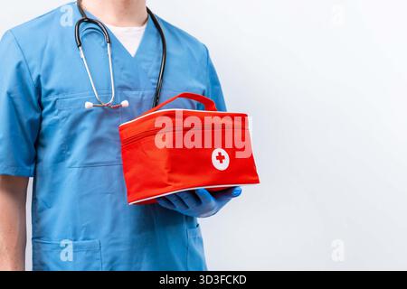 Un medico di sesso maschile in uniforme blu sta con una borsa medica di pronto soccorso rossa su uno sfondo isolato Foto Stock