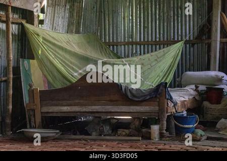 Vista interna di una casa rurale molto disordinata e povera in Bangladesh, caratterizzata da un semplice e vecchio letto in legno con zanzariera protettiva (o Foto Stock