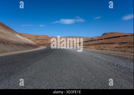 Ripresa da un angolo basso di una strada che attraversa il deserto marocchino sotto un cielo blu vivace con nuvole bianche sparse Foto Stock