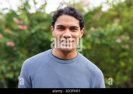 Uomo in giardino con maglione in maglia grigio-blu e fotocamera sorridente con fiori rosa Foto Stock