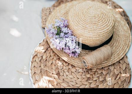 Elegante letto piatto con cappello di paglia decorato con fiori lilla e spago posizionato su un tappeto in tessuto naturale. La luce soffusa e i toni neutri creano calma e somme Foto Stock
