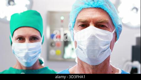 Chirurgo anziano in piedi che indossa un cappellino, un camice e una maschera in sala operatoria, con luci e pannelli Foto Stock