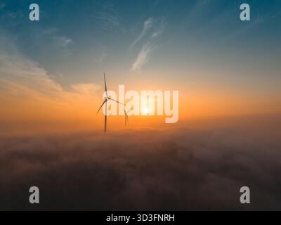 Turbine eoliche che si innalzano al di sopra di un paesaggio nebbioso all'alba con una luce calda Foto Stock